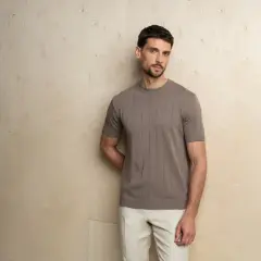 Mężczyzna prezentuje elegancki, casualowy styl. Brązowa, prążkowana koszulka z krótkim rękawem i beżowe spodnie tworzą harmonijną całość.