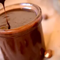 Un chorrito de crema de chocolate cae en un frasco lleno de esta delicia. Avellanas y tabletas de chocolate completan la escena.