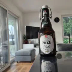 Eine Flasche Moritz Fiege Pils steht auf einem Tisch. Im Hintergrund ein Wohnzimmer mit Sofa und Fernseher.