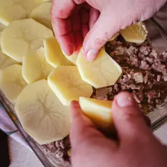 Ręce układają plastry ziemniaków na warstwie mięsa mielonego, tworząc pyszną zapiekankę ziemniaczaną z mięsem mielonym.