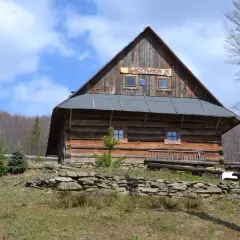Drewniana bacówka w górach z napisem "BACÓWKA" na dachu, otoczona drzewami i kamiennym murkiem.