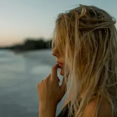 Kobieta z blond włosami rozjaśnionymi przy twarzy, na tle plaży i morza.