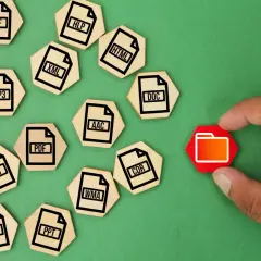 Un dedo toca una carpeta roja, rodeada de fichas de madera con extensiones de archivo como ZIP, MP3, DOC, etc. que representan un archivo comprimido.