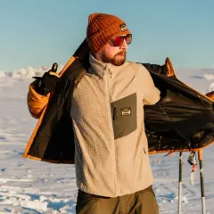 Man i orange mössa och solglasögon visar upp sin lager på lager-klädsel i snölandskap med skidstavar.