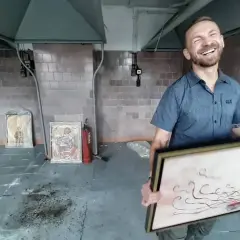 Uśmiechnięty kossakowski malarz prezentuje swoje dzieła. W tle widać obrazy i gaśnicę.