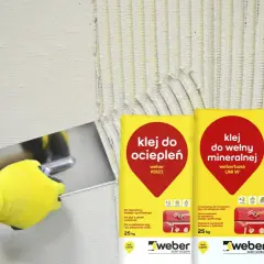 Ręka w żółtej rękawicy nakłada klej do wełny mineralnej na ścianę. Obok worki z klejem.