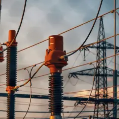 Trzy izolatory z brązowymi głowicami, przygotowane do dystrybucji energii. W tle widać wieżę przesyłową, symbolizującą napięcie fazowe a międzyfazowe.