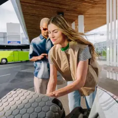 Para podróżuje na wakacje, pakując bagaż do samochodu. W tle widać zielony autokar, który może zabrać ich do Kołobrzegu lub Mielna.