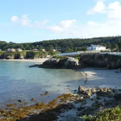 La **playa Burela** se asoma entre rocas y acantilados, con casitas blancas coronando la colina. El mar turquesa acaricia la arena dorada donde pasean algunas personas.