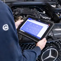 Meccanico esegue la prima revisione auto con tablet diagnostico davanti al motore di una Mercedes.