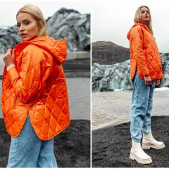 Pomarańczowa, pikowana kurtka jesienna oversize damska z kapturem, zestawiona z jeansami i białymi botkami, na tle surowego krajobrazu.