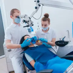 Dentysta pod mikroskopem wykonuje ekstyrpację miazgi u pacjentki. Asystentka pomaga.