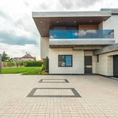 Nowoczesny dom z podjazdem z kostki brukowej. W oddali widać drzewa i inne domy. Lampa ogrodowa słupek dyskretnie oświetla ścieżkę.