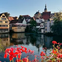 Blick auf Bamberg mit Fachwerkhäusern am Wasser. Rote Geranien blühen im Vordergrund. Entdecken Sie Bamberg Geheimtipps entlang des Flusses.