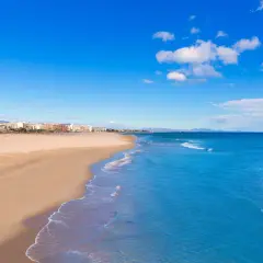 Amplia playa de arena dorada en Sagunto, con el mar Mediterráneo azul y edificios de la costa al fondo bajo un cielo despejado.
