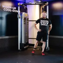 Mężczyzna w czarnej koszulce "Fabryka Siły" wykonuje ćwiczenie na maszynie typu cable cross. Z pomocą taśmy na kostce próbuje **pull through** z boku, wzmacniając mięśnie pośladkowe.
