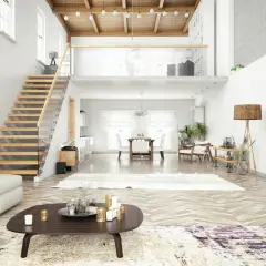 Un grand salon lumineux avec un escalier en bois menant à une mezzanine sous toit en pente. Un canapé blanc, une table basse et une salle à manger sont visibles.
