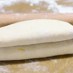 Ciasto francuskie złożone na pół, gotowe do dalszej obróbki. Wałek i mąka na blacie sugerują, jak zrobić ciasto francuskie.