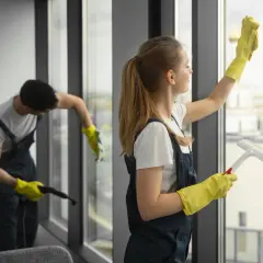 Dwie osoby w żółtych rękawiczkach myją okna. Jedna używa ściągaczki, druga parownicy.