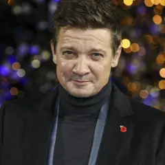 Jeremy Renner, med en vallmo på kavajen, ser allvarlig ut mot en bakgrund av julbelysning. Efter sin svåra olycka är han tillbaka.
