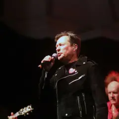 Kawalec śpiewa, a budka suflera czuwa. Muzycy na scenie, w tle gitara i czerwona koszula.