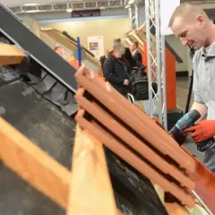 Dekarz w czerwonych rękawicach używa wkrętarki do montażu dachówek na konstrukcji dachu.