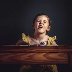 Nerwowe dziecko 2 lata, zaciśnięte usta, oczy zamknięte, siedzi przy drewnianym biurku z kredkami.