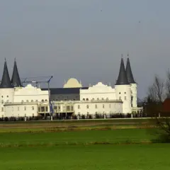 Biały budynek przypominający zamek z ciemnymi wieżami, w tle żurawie budowlane.