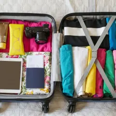 Packar handbagage på flyg. En kamera, solkräm, glasögon, surfplatta, pass och färgglada kläder ligger prydligt i resväskan.