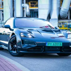 Elegancki Porsche Taycan w ciemnym kolorze, zaparkowany przed industrialnym budynkiem z rurami.