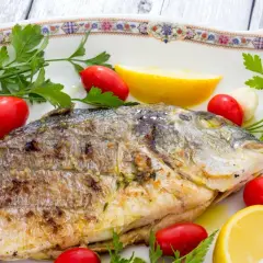 Orata alla griglia dorata e succulenta, guarnita con pomodorini, spicchi d'aglio, limone e prezzemolo fresco.