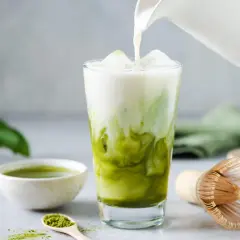 Milch wird in ein Glas mit Eis und Matcha gegossen. Ein Löffel mit Matcha-Pulver und eine Schale mit Matcha-Tee sind daneben zu sehen. Ein traditioneller Matcha-Besen liegt auf dem Tisch.