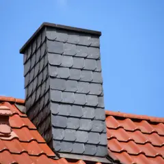 Moderner gemauerter Schornstein verkleidet mit grauen Schieferplatten, überragt ein rotes Ziegeldach unter blauem Himmel.