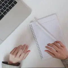 Ręce piszą notatki do CV po niemiecku przy laptopie, obok filiżanka kawy.