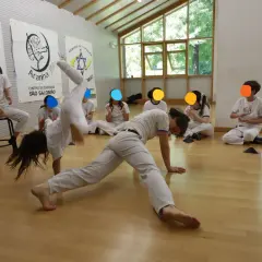 Bambini praticano capoeira in una palestra luminosa. Uno fa una capriola, altri osservano e applaudono.