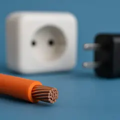 Pomarańczowy kabel z odsłoniętymi miedzianymi żyłami, białe gniazdko i czarna wtyczka. Czy plastik przewodzi prąd? Nie, izoluje.