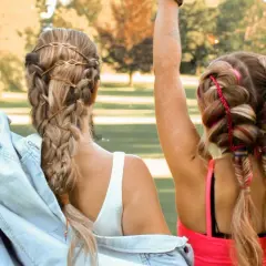 Zwei Mädchen mit geflochtenen Haaren, die sich für festival frisuren einfach eignen. Eine trägt braune Bänder, die andere bunte Fäden.