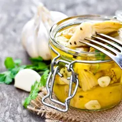 Deliziosi carciofini sott'olio fatti in casa, conservati in un barattolo di vetro con aglio e prezzemolo. Un forchettone ne solleva uno, pronto per essere gustato.