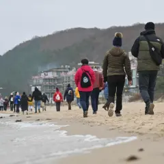Spacer po plaży, gdy jest długi weekend. Ludzie w ciepłych ubraniach idą brzegiem morza, w tle widać zabudowania i wzgórza.
