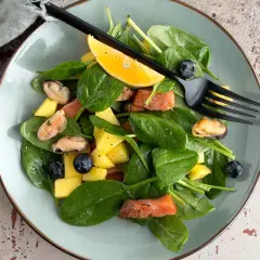 Insalata con mango, salmone, cozze e mirtilli, guarnita con spicchio d'arancia e forchetta nera.