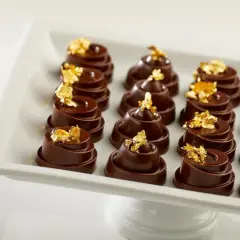Douze chocolats fins, une composition alimentaire luxueuse, ornés de feuilles d'or comestibles, présentés sur un plat blanc.
