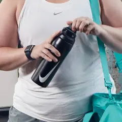 Osoba w białej koszulce sportowej trzyma czarną butelkę Nike. Obok wisi turkusowa torba.