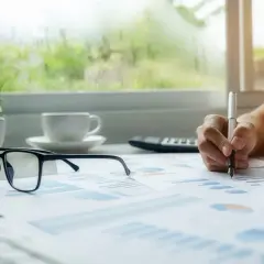 Okulary, filiżanka kawy i ręka pisząca piórem na wykresach na biurku.