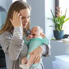 Erschöpfte Mutter hält weinendes Baby. Der 12 wochen schub macht ihr zu schaffen. Sie hält sich die Stirn.