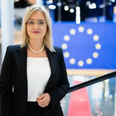Magda Adamowicz, w eleganckim stroju, stoi przed flagą Unii Europejskiej. Jej wiek nie jest widoczny, ale emanuje pewnością siebie.