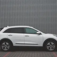 Biały Kia Niro EV stoi na tle szarej, falistej ściany. Elegancki design i nowoczesne linie podkreślają jego ekologiczny charakter.