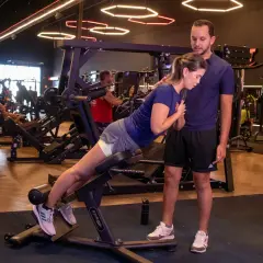En kvinna utför 45 graders hyperextension med en tränare som ger stöd. Träningspasset sker på ett modernt gym.