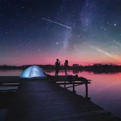 Para na molo pod rozgwieżdżonym niebem, obok namiotu. Woda odbija barwy zachodzącego słońca i gwiazdy na niebie.