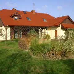 Dom z czerwonym dachem naczółkowym, z werandą i ogrodem pełnym traw ozdobnych.