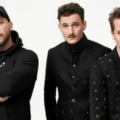 Trzech mężczyzn w czarnych ubraniach, jeden w czapce z daszkiem, na białym tle.